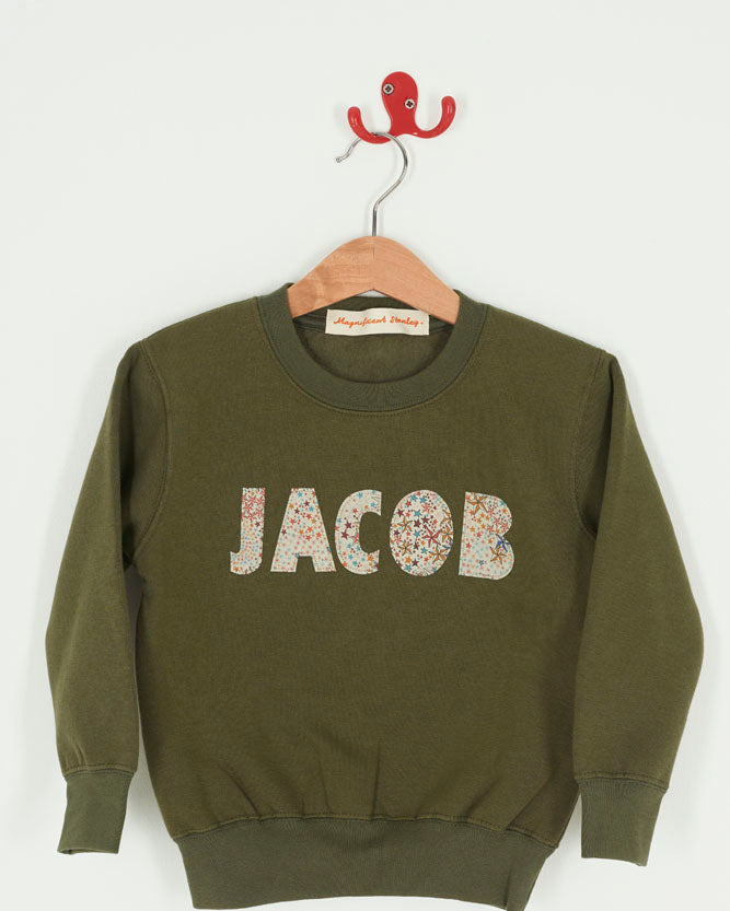 Green sweatshirt with 'JACOB' sewn on it in Liberty star Adelajda fabric, hanging on a wooden hanger against a white background.