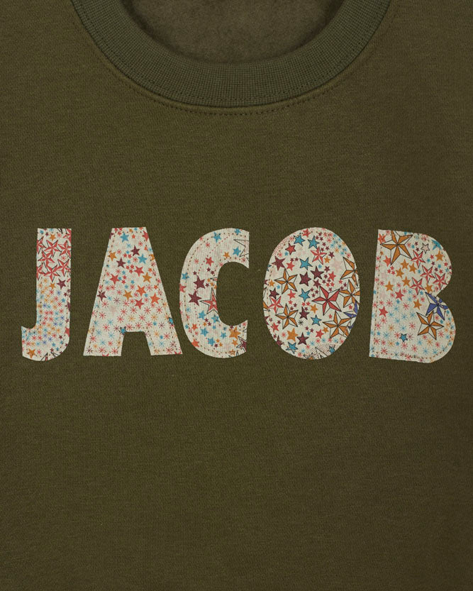 Green sweatshirt with 'JACOB' sewn on it in Liberty star Adelajda fabric, hanging on a wooden hanger against a white background.