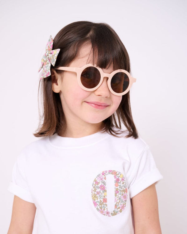A Child wearing a white t-shirt with a small floral liberty print initial on the front.
