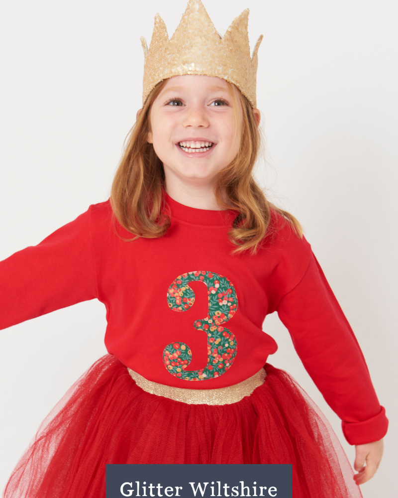 Child wearing a red t-shirt with the number 3 in liberty Wiltshire print, gold crown, and red skirt on a white background.