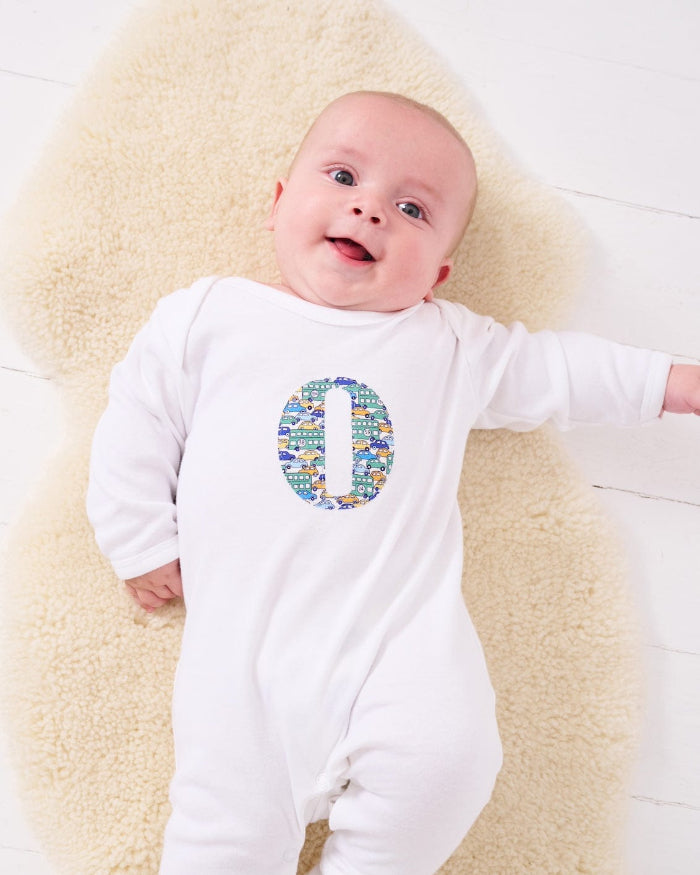 a happy baby wearing a personalised cotton baby romper with his initial on the front in hop on hop off liberty print featuring cars and London buses.