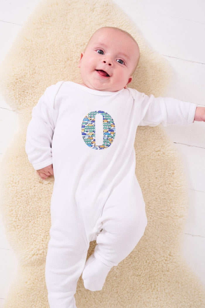 a happy baby wearing a personalised cotton baby romper with his initial on the front in hop on hop off liberty print featuring cars and London buses.