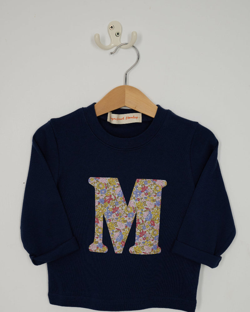 A navy blue long-sleeve t-shirt hanging on a wooden hanger with a white wall hook behind it. The sweatshirt features a large capital letter 'M' on the front filled with a Liberty print floral cotton