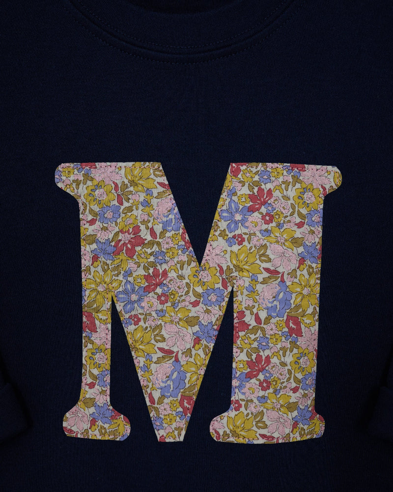 A close-up of a navy blue kids t-shirt featuring a large letter 'M' graphic filled with a Liberty print floral Juila Belle pattern.