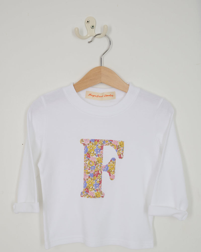 A white long-sleeve t-shirt with a large Juila Belle Liberty floral-patterned letter 'F' on the front, hanging on a wooden hanger against a plain off-white wall.