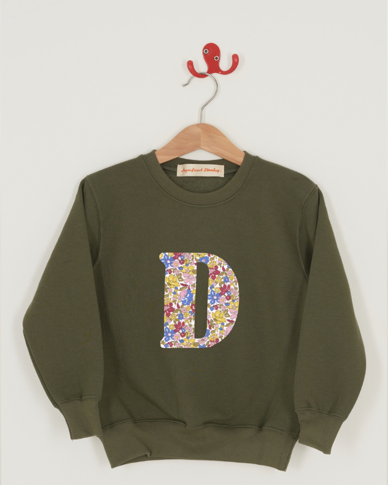 An olive green crewneck sweatshirt hanging on a wooden hanger with a red hook, featuring a large Liberty print floral-patterned letter 'D' on the front. The fabric name 'Julia Belle' is printed below the sweatshirt.