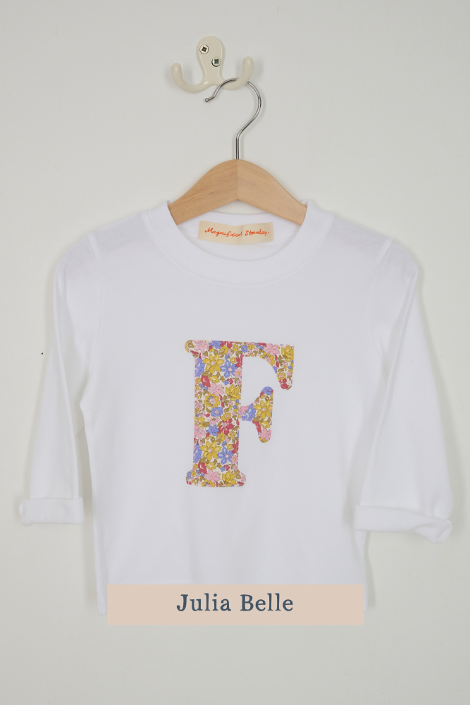 A white long-sleeve t-shirt with a large floral-patterned letter 'F' on the front, hanging on a wooden hanger against a light wall. A beige label at the bottom reads 'Julia Belle'.