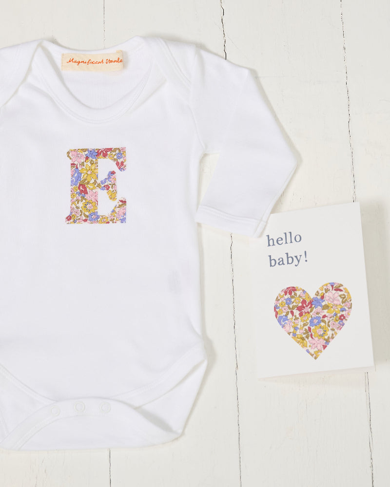 A white long-sleeve baby bodysuit with a colourful floral letter 'E' on the chest, displayed next to a matching 'hello baby!' Liberty fabric card on a white wood background.