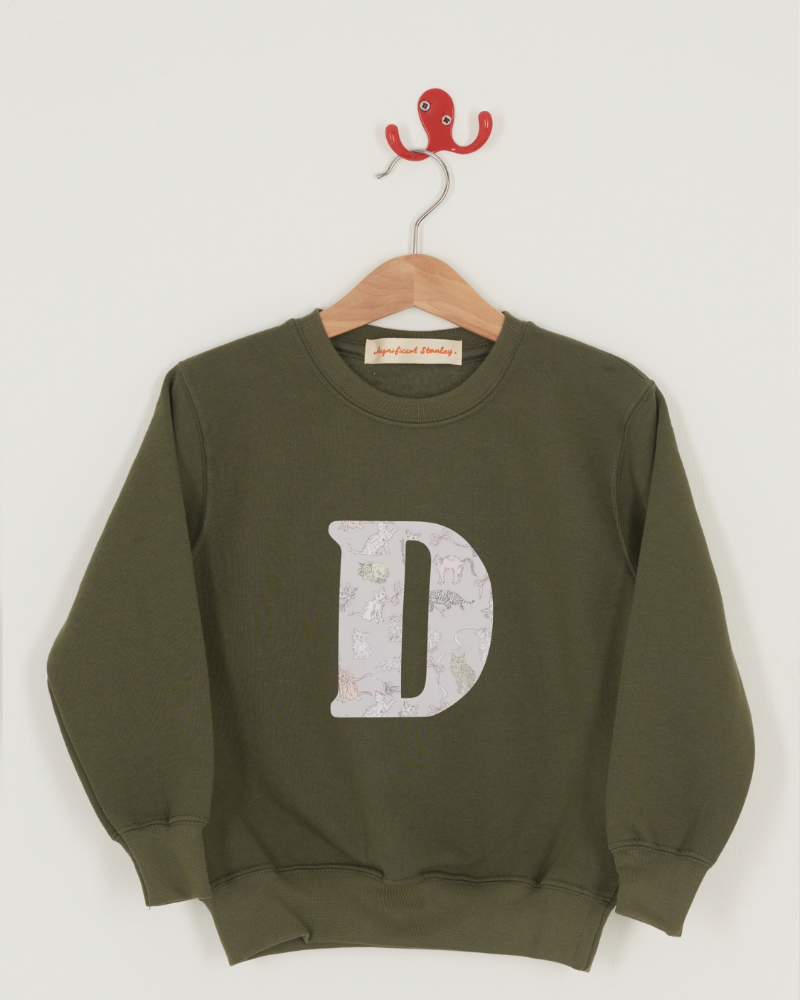 A khaki green crewneck sweatshirt hanging on a wooden hanger with a red hook, featuring a large Liberty print cat patterned letter 'D' on the front. The fabric name 'Koh & Eito' is printed below the sweatshirt.