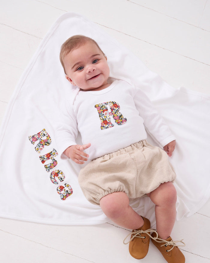 A newborn baby girl wearing a Personalised baby Bodysuit in Libby floral Liberty Print. She is laying on a personalised cotton baby blanket with her name sewn on.