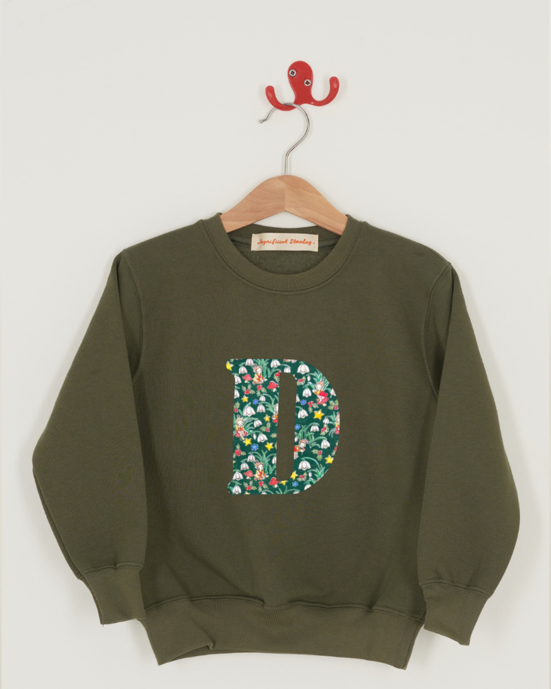 An olive green crewneck sweatshirt hanging on a wooden hanger . The sweatshirt features a large Liberty print Elves letter 'D' on the front filled with a colourful floral pattern featuring snowdrops, stars and holly.