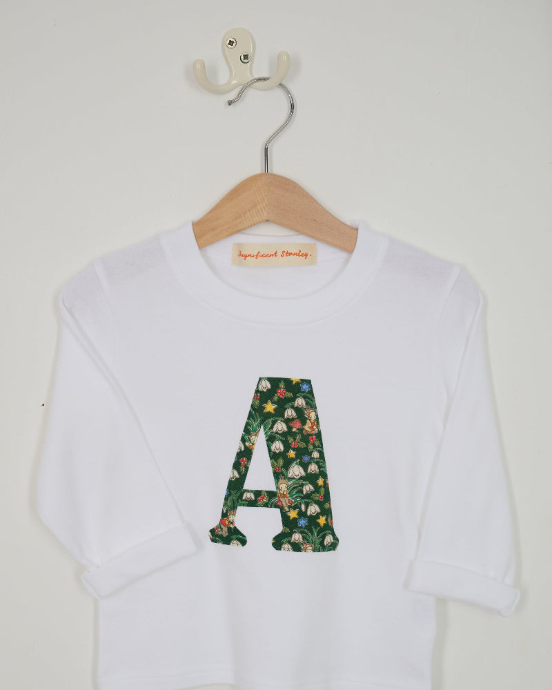 A white long-sleeve top for kids, featuring a large embroidered letter 'A' on the front filled with a green floral Liberty Elves pattern. The shirt is displayed on a wooden hanger with a white wall hook behind it.