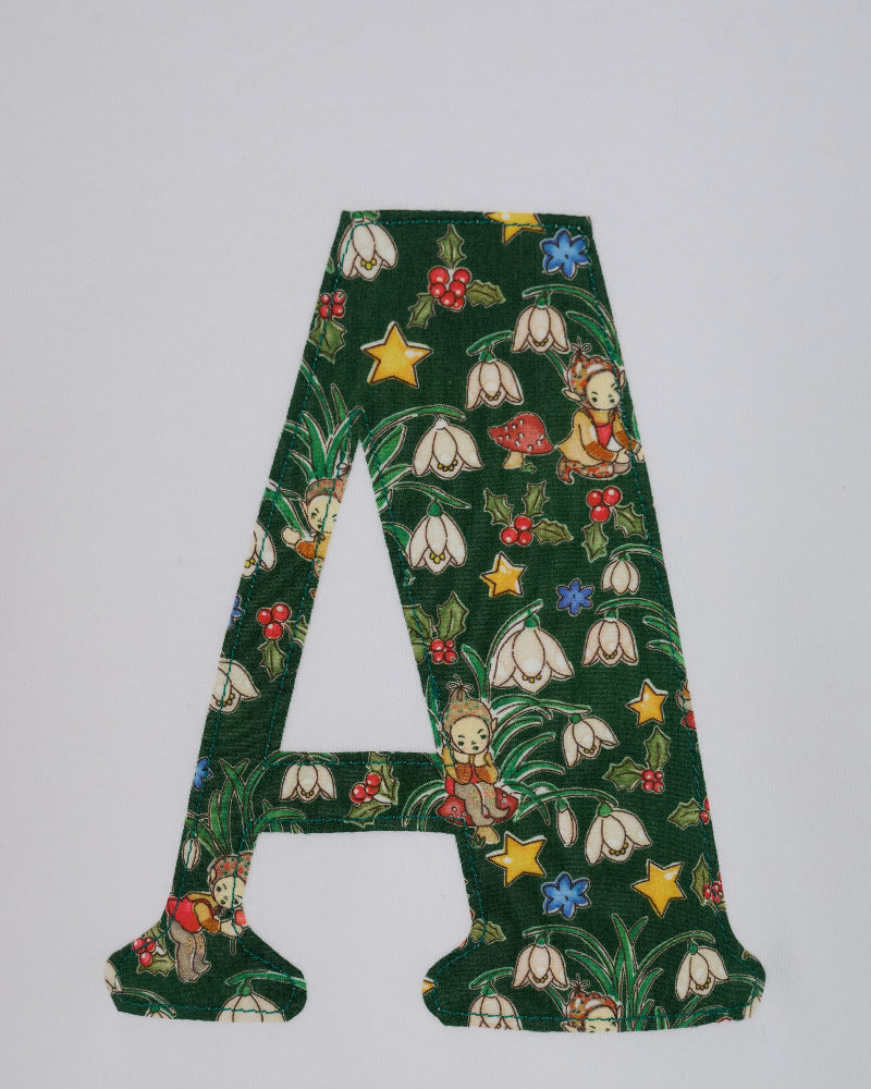 A decorative capital letter A cutout featuring a Liberty Elves pattern with white snowdrops, yellow stars, red berries, and a small elf on a dark green background, shown against a plain light gray backdrop.