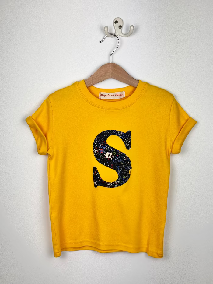 Yellow t-shirt with a large black letter 'S' on a hanger against a white background