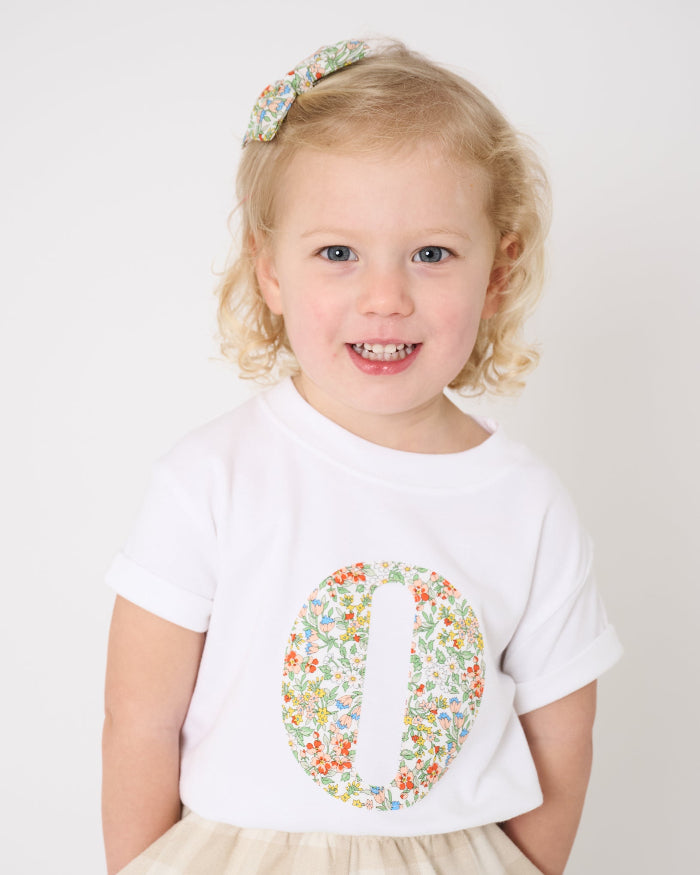 A 2 year old girl wearing a summery personalised white cotton t-shirt with her initial cut from Nazanin floral liberty fabric.