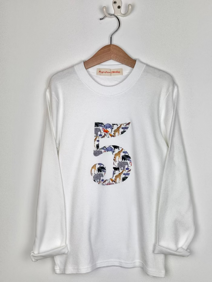 A white kids birthday t-shirt hanging against a white wall. The t-shirt features a number 5 cut from liberty zoo print.