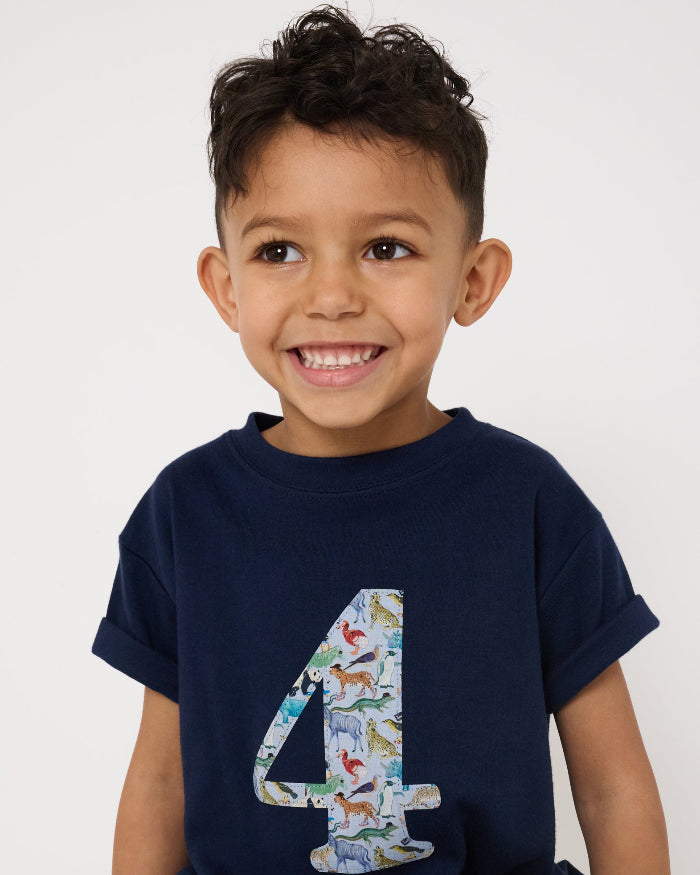 A 4 year old boy wearing a navy cotton t-shirt with the number 4 sewn on the front cut from liberty Quey 2 zoo animal print.