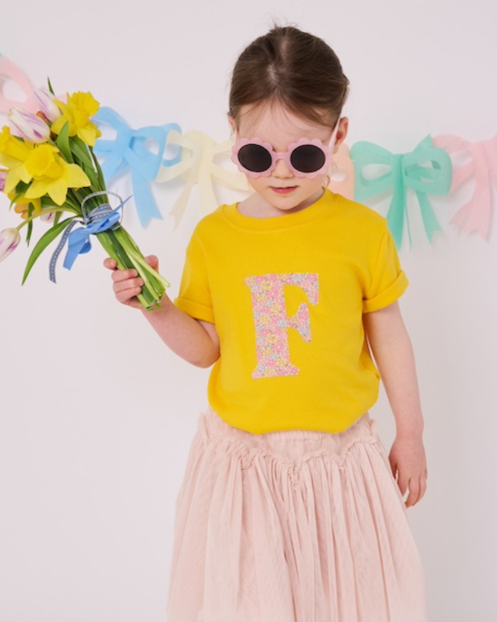 a smiling 3 year old girl wearing a yellow personalised t-shirt with her initial cut from floral liberty print on the front.