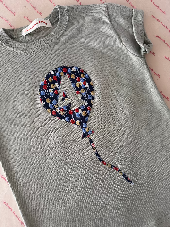 A sage green short-sleeve kids t-shirt featuring a balloon applique made of colourful ballons Liberty print. The shirt is laid flat on a pink patterned surface, showing the brand label 'Magnificent Stanley'.
