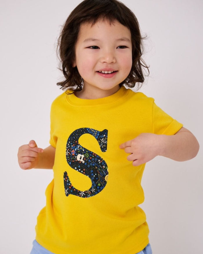 a smiling 3 year old boy wearing a personalised kids yellow t-shirt with an initial cut from adelaide in space liberty print on the front