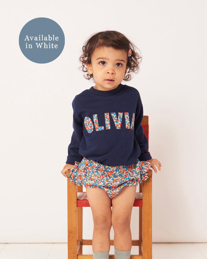 Child wearing a navy sweatshirt with 'OLIVIA' printed on it, sitting on a chair against a white background.