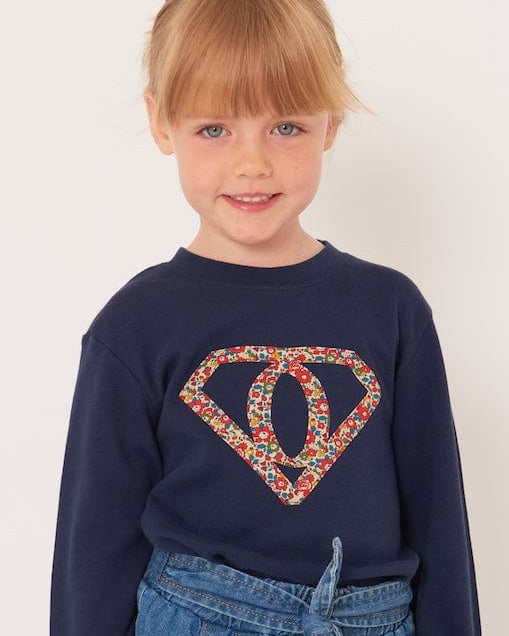 A 5 year old girl wearing a personalised navy t-shirt with a superhero motif initial made from floral Liberty print fabric.