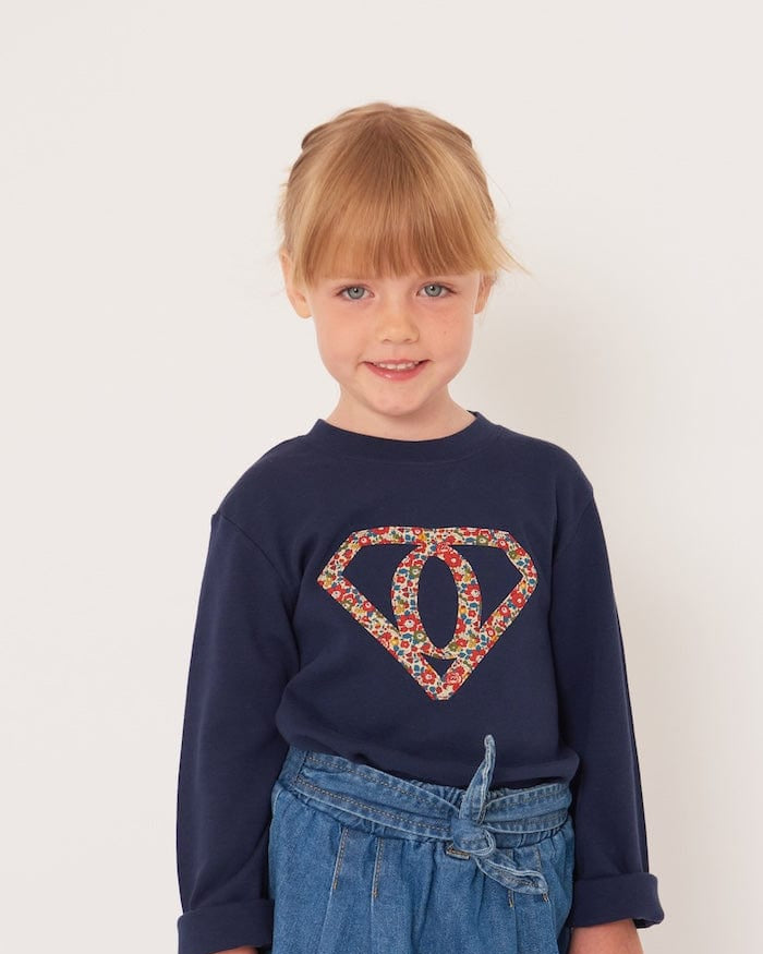 A 5 year old girl wearing a personalised navy t-shirt with a superhero motif initial made from floral Liberty print fabric.