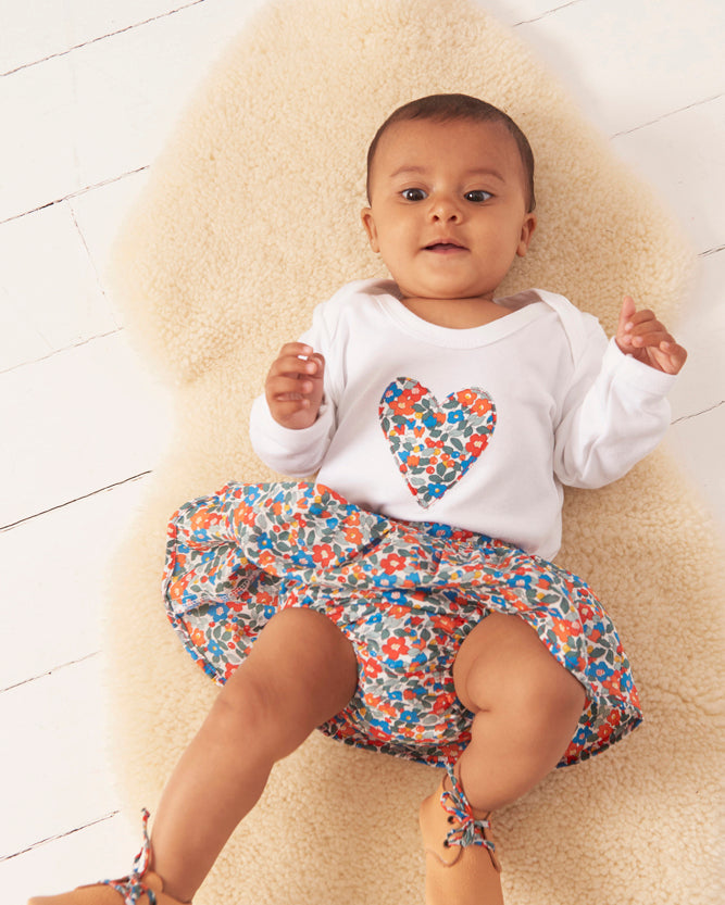 Baby wearing a white bodysuit with a colourful liberty print heart design and a floral skirt on a light wooden floor.