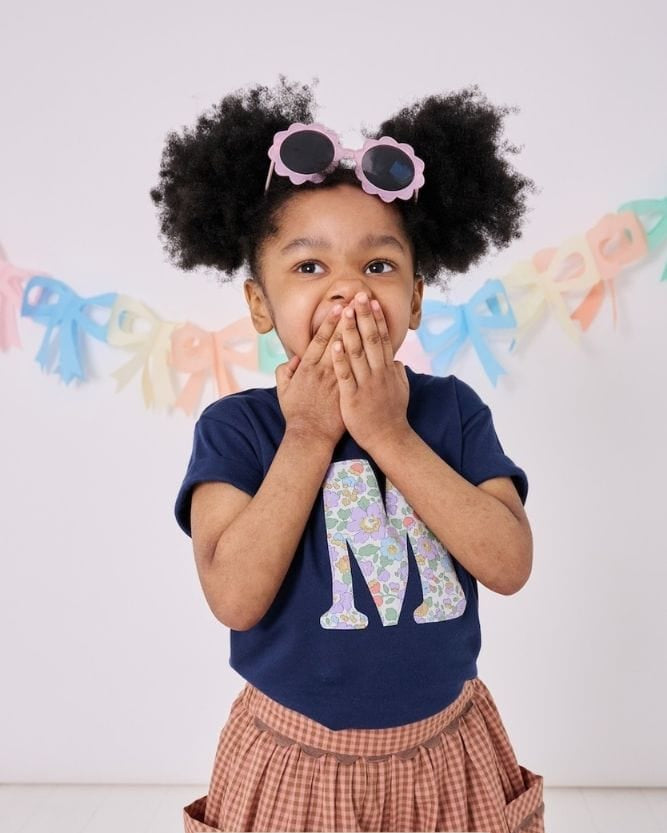 a 3 year old boy wearing a personalised kids navy t-shirt with an initial cut from betsy purple floral liberty print on the front.