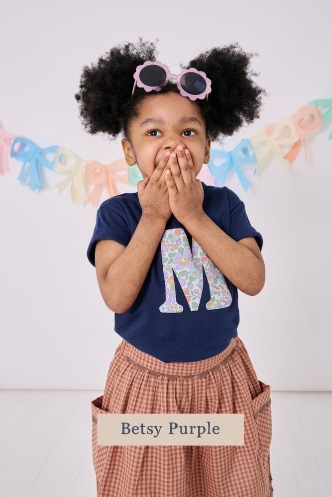 a 3 year old boy wearing a personalised kids navy t-shirt with an initial cut from betsy purple floral liberty print on the front.