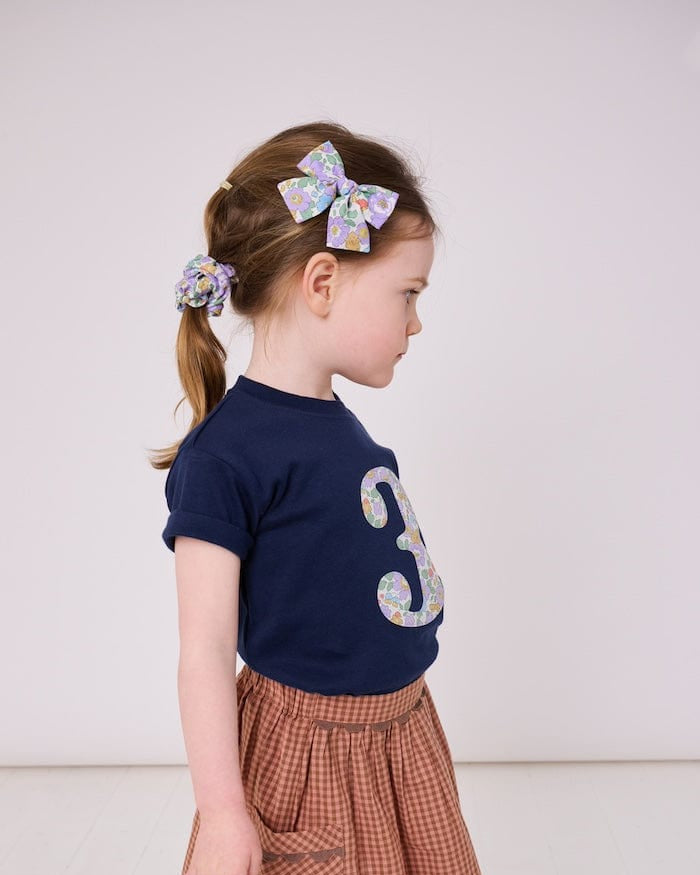 a 3 year old girl wearing a navy birthday t-shirt with a number 3 cut from liberty betsy purple floral print on the front and a matching floral scrunchie.