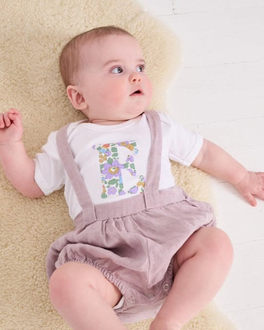 A smiling newborn baby wearing a personalised white bodysuit with an initial on the front cut from betsy purple Liberty floral fabric.