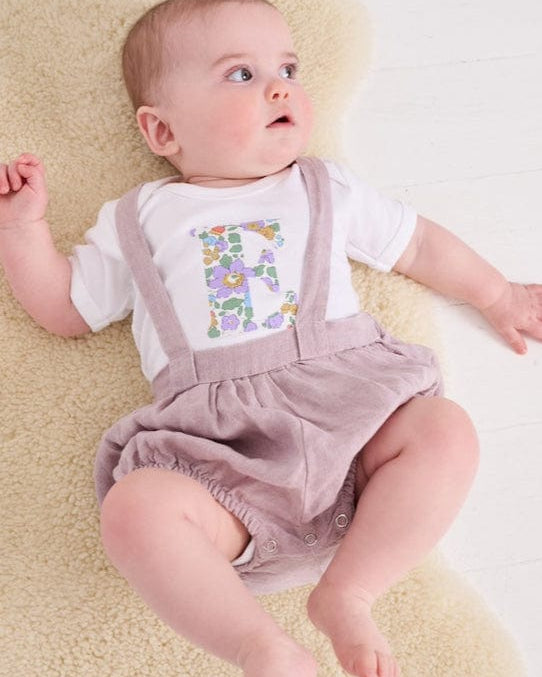 A smiling newborn baby wearing a personalised white bodysuit with an initial on the front cut from betsy purple Liberty floral fabric.