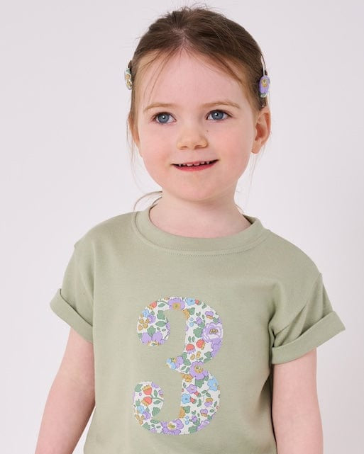a 3 year old girl wearing a birthday number 3 kids sage green t-shirt with a number cut from betsy purple liberty print on the front.