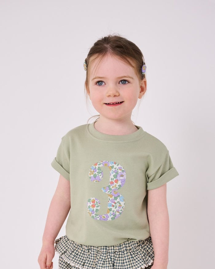 a 3 year old girl wearing a birthday number 3 kids sage green t-shirt with a number cut from betsy purple liberty print on the front.