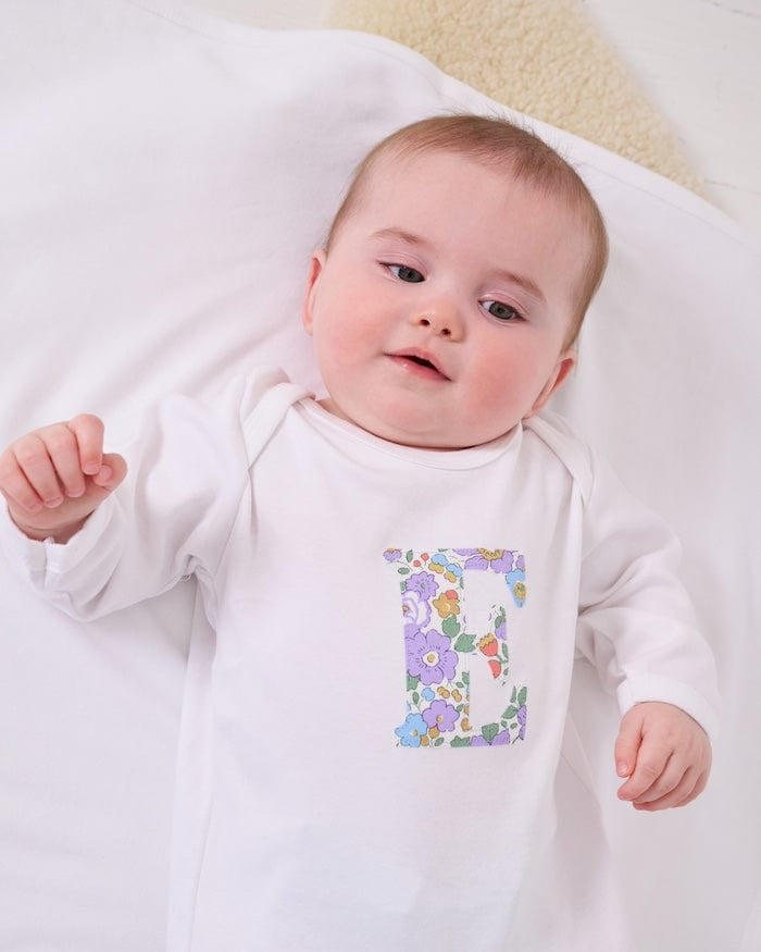 a happy baby wearing a cotton baby romper with her initial on the front in betsy purple floral liberty print.