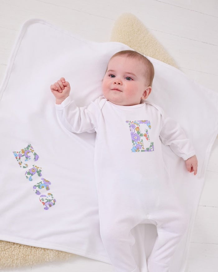 a happy baby wearing a cotton baby romper with her initial on the front in betsy purple floral liberty print. she is laying on a  cotton blanket with her name sewn on.