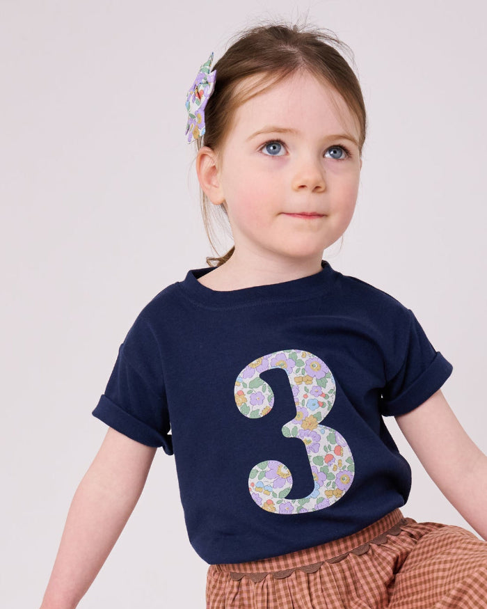 A 3 year old birthday girl wearing a navy cotton kids birthday t-shirt with the number 3 in Betsy purple floral Liberty print on the front.