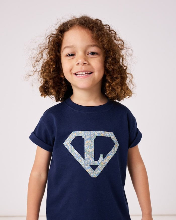 a 4 year old boy wearing a personalised navy t-shirt with his initial inside a superhero emblem made from Liberty print