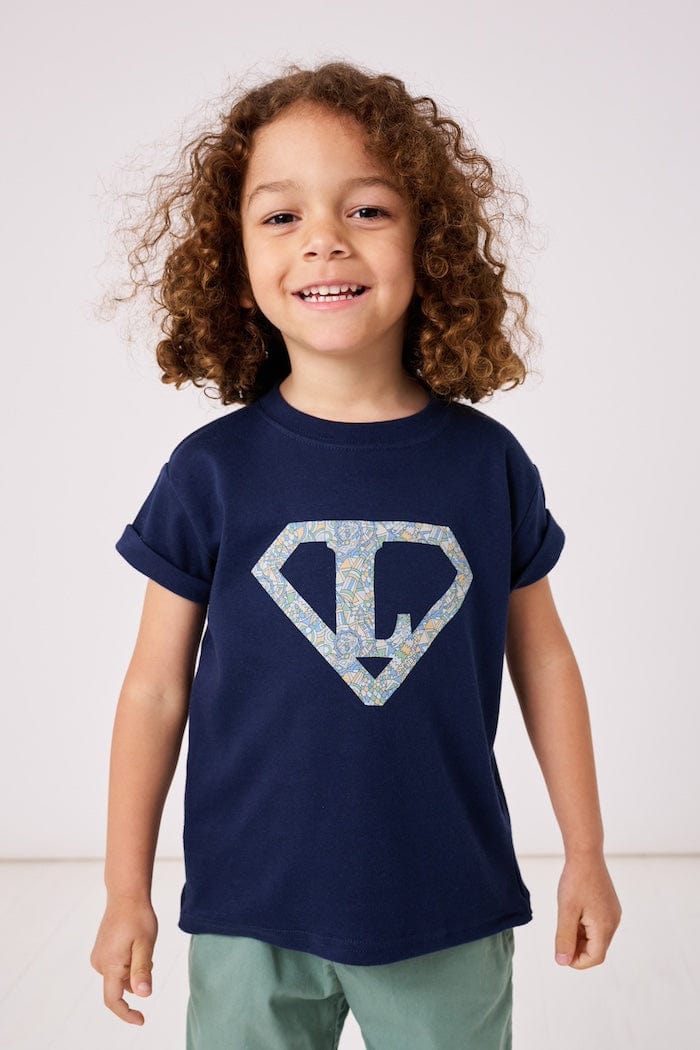 a 4 year old boy wearing a personalised navy t-shirt with his initial inside a superhero emblem made from Liberty print