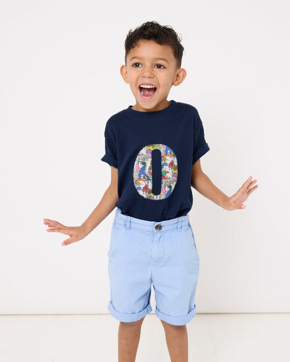 A 4 year old boy wearing a bespoke navy t-shirt with a colourful letter initial design in Liberty dinosaur print and light blue shorts on a white background