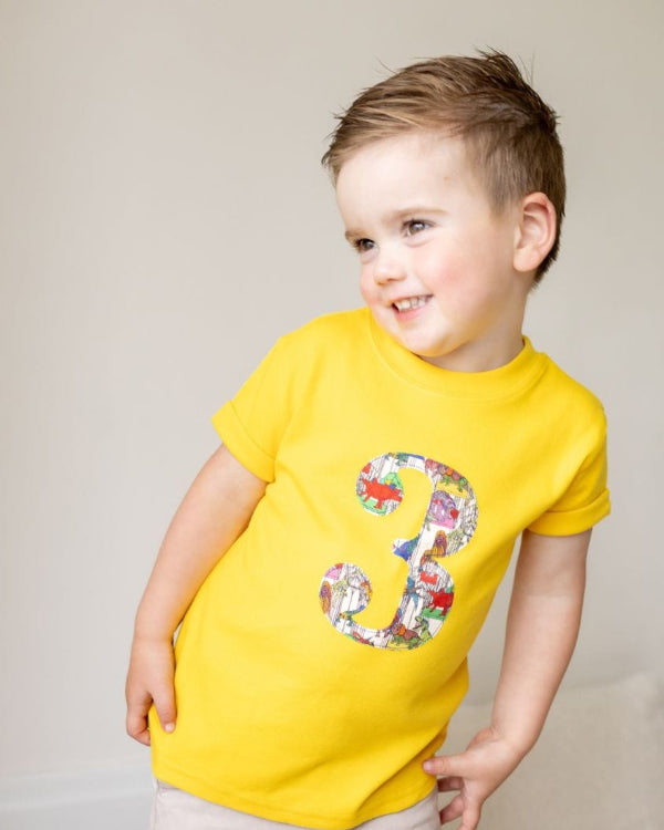 a smiling 3 year old boy wearing a yellow birthday t-shirt with a number 3 cut from claws roars n' dinosaurs liberty print on the front.