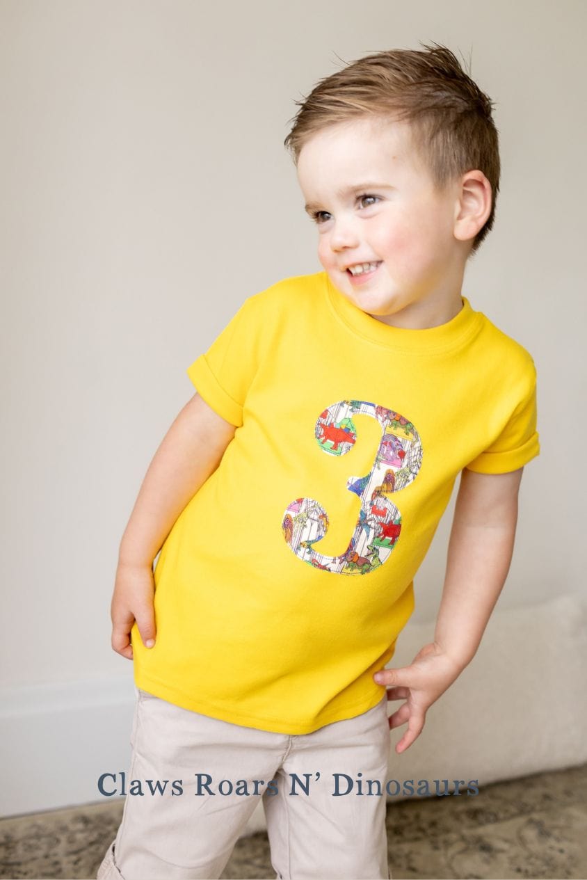 a smiling 3 year old boy wearing a yellow birthday t-shirt with a number 3 cut from claws roars n' dinosaurs liberty print on the front.