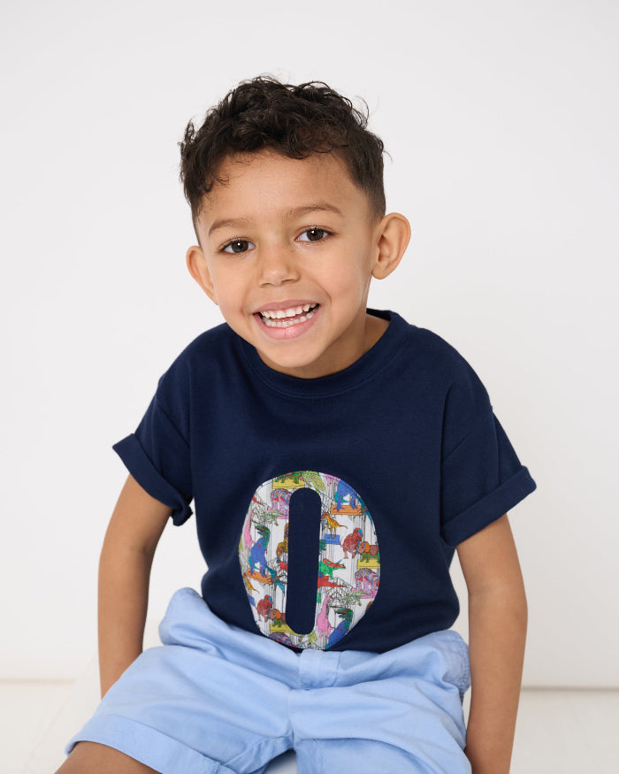 A 4 year old boy wearing a personalised navy t-shirt with a colourful letter design in Liberty dinosaur print and light blue shorts on a white background