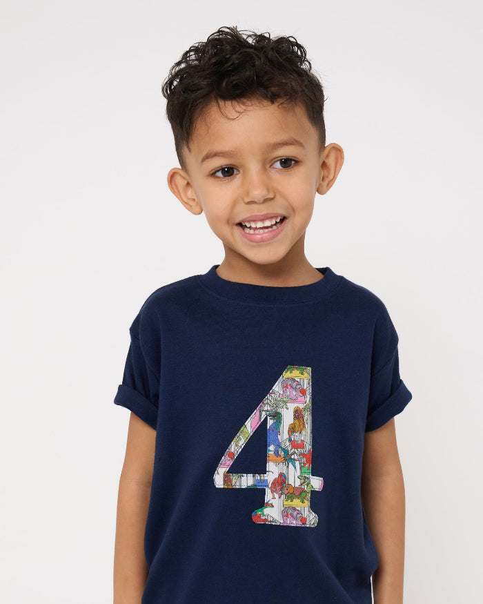 A 4 year old boy wearing a navy t-shirt with a colourful number 4 design in Liberty dinosaur print and light blue shorts on a white background