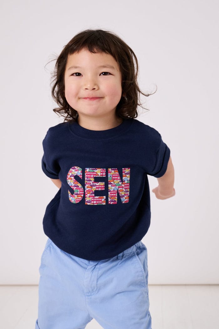 A smiling 3 year old boy wearing a navy cotton personalised t-shirt with his name on cut from liberty print featuring London buses and vintage cars.