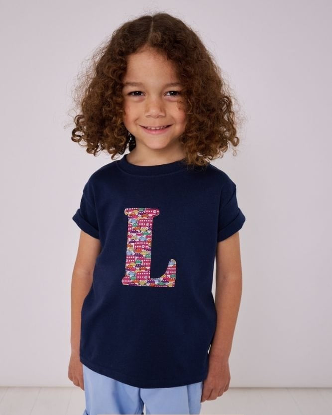 a 5 year old boy wearing a personalised kids navy t-shirt with an initial cut from hop on hop off liberty print featuring london buses and vintage cars
