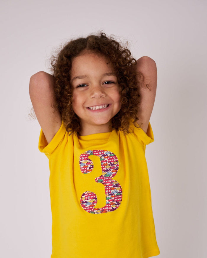 a 3 year old birthday boy wearing a yellow t-shirt with a number 3 cut from Hop on hop off red cars and london bus liberty print on the front.