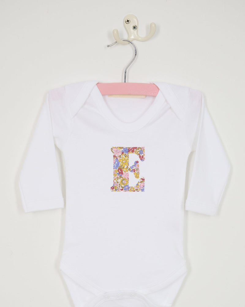 A white long-sleeve baby bodysuit laid flat on a yellow and pink sparkly Liberty Julia Belle floral fabric background. The bodysuit has a decorative floral block-letter 'E' on the chest.