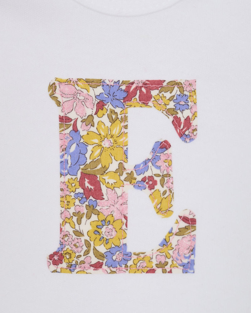 A close up of a white long-sleeve baby bodysuit laid flat on a yellow and pink sparkly Liberty Julia Belle floral fabric background. The bodysuit has a decorative floral block-letter 'E' on the chest.